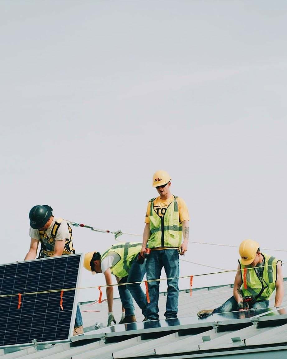 Four JR&CO crew members in hard hats and hi-vis vests installing solar panels on a metal roof
