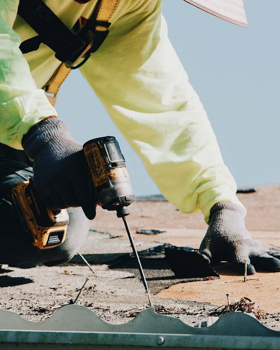 JR&CO roofer in a hi-vis yellow shirt fastening roof decking with a cordless drill under a blue sky